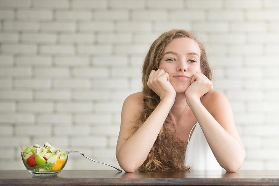 devojka nalakćena na stolu pored nje činija sa voćem i povrćem - ortoreksija