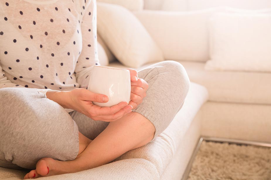 Mlađa žena u pidžami prekrštenih nogu drži šolju čaja