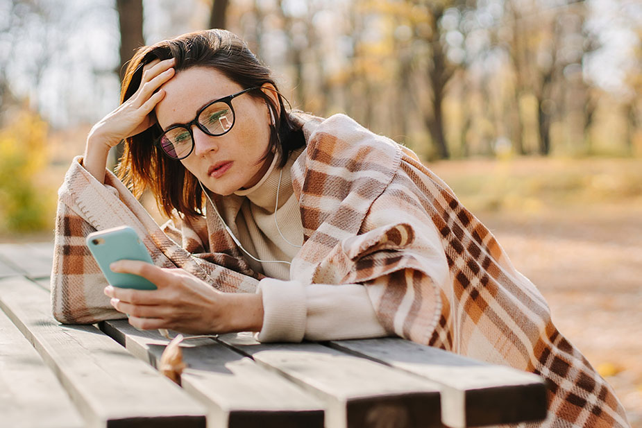 Mlađa žena sedi na klupi u prirodi ogrnuta ćebetom i gleda u telefon