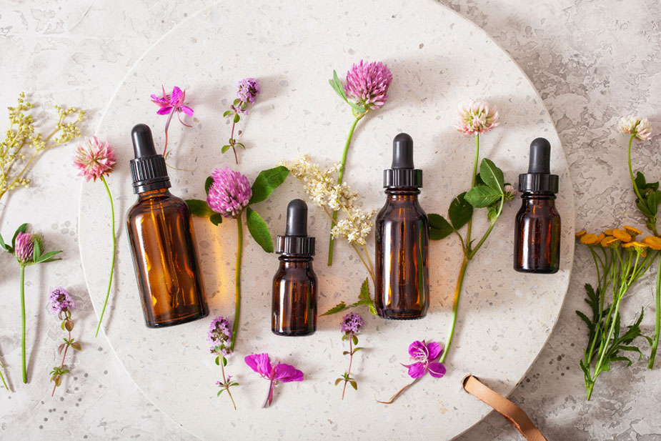 Raznolike boce esencijalnih ulja okružene svežim cvetovima na mermernom stolu.