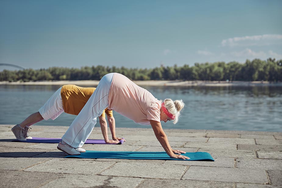 Starija žena u sportskoj odeći radi pozu planinara na joga podlozi pored jezera na sunčan dan - fitnes program.