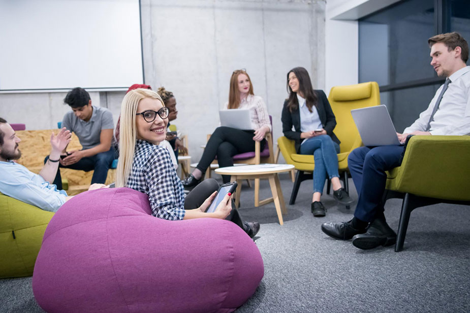 Ljudi sede u udobnim stolicama i vrećama za sedenje, razgovaraju i koriste laptopove, prikazujući opuštenu atmosferu na poslu.