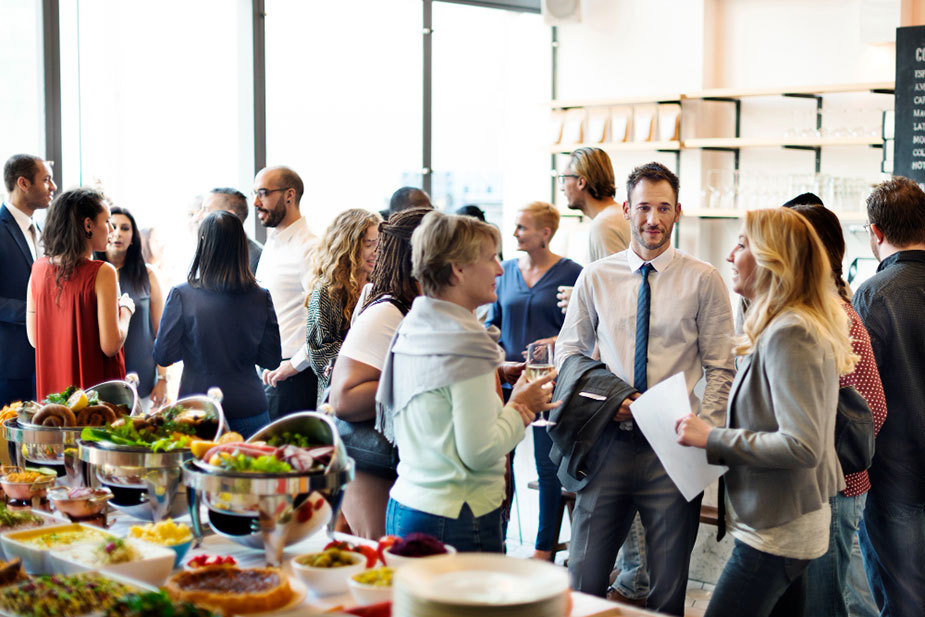 Grupa ljudi stoji i razgovara na poslovnom događaju uz sto sa hranom, naglašavajući važnost umrežavanja i neformalnih susreta u poslovnom svetu.