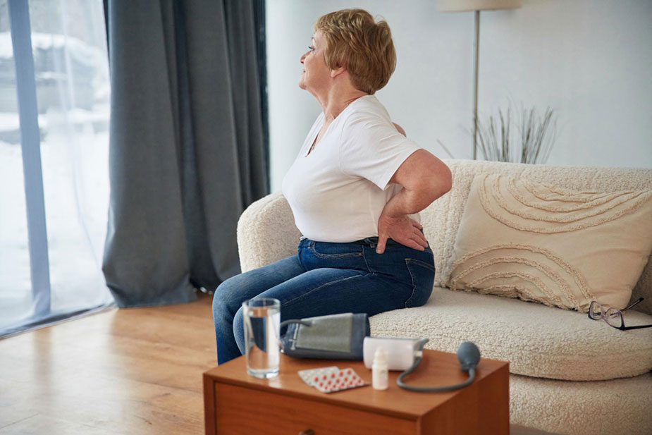 Starija dama sedi na trosedu i drži se za leđa. Ispred nje je sto sa lekovima.
