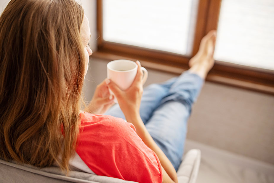 Žena sedi uz prozor i pije kafu u miru – simbol smirenog početka dana i prevencije stresa.