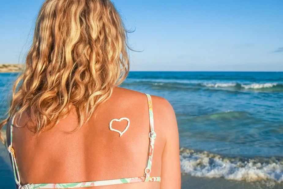 Žena sa kovrdžavom kosom na leđima, sa oblikom srca od kreme za sunčanje na leđima, na plaži.