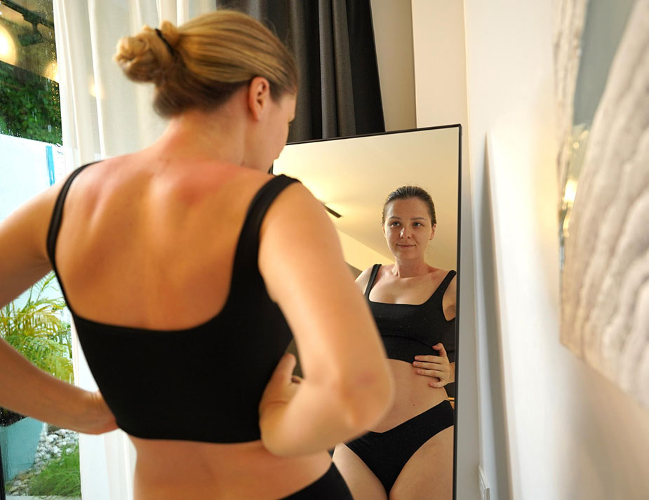 Žena posmatra svoj stomak u ogledalu, primećujući povećanje telesne težine.