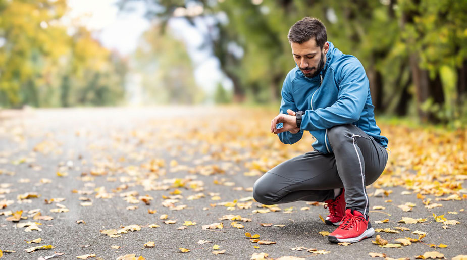 Muškarac u sportskoj garderobi čuči na stazi prekrivenoj lišćem i gleda u sportski sat.